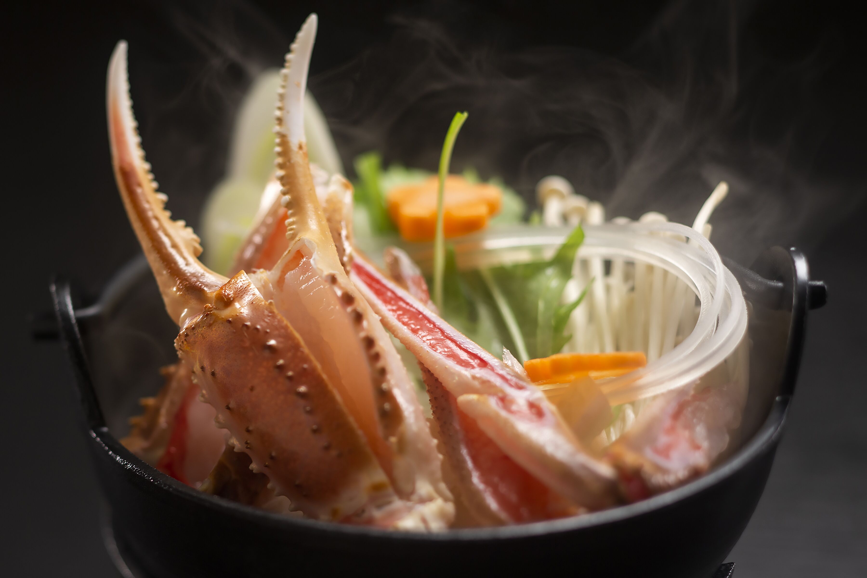 A lobster in a black bowl with a steam effect, surrounded by vegetables.