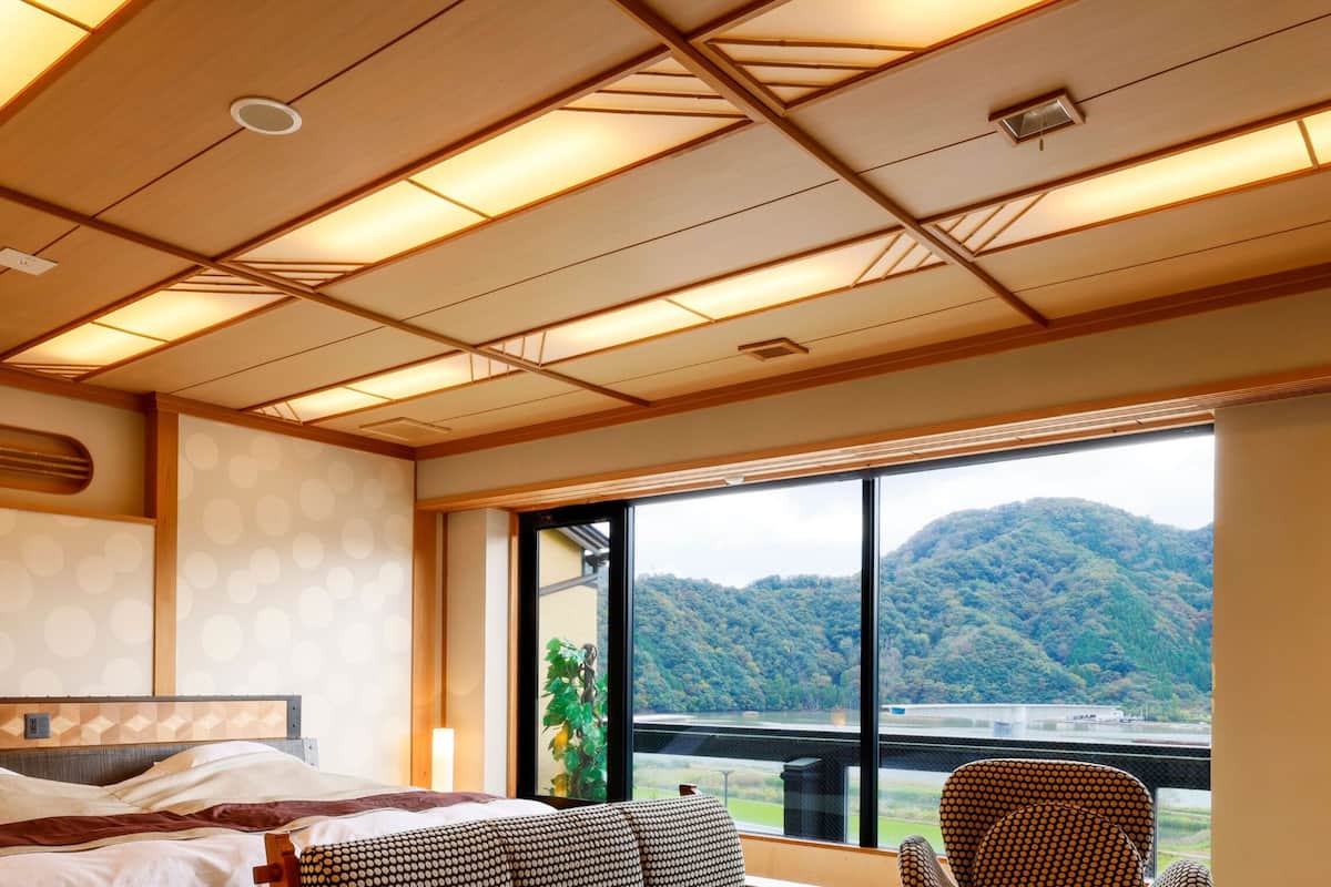Ein Hotelzimmer mit einem großen Bett, einem Fenster mit Blick auf einen Berg und einen See, sowie einer Decke mit Holzbalken und eingelassener Beleuchtung.