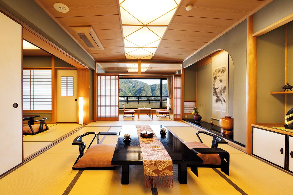 Ein traditioneller japanischer Raum mit Tatami-Böden, einem niedrigen Tisch mit Kissen und Blick auf Berge durch große Fenster.