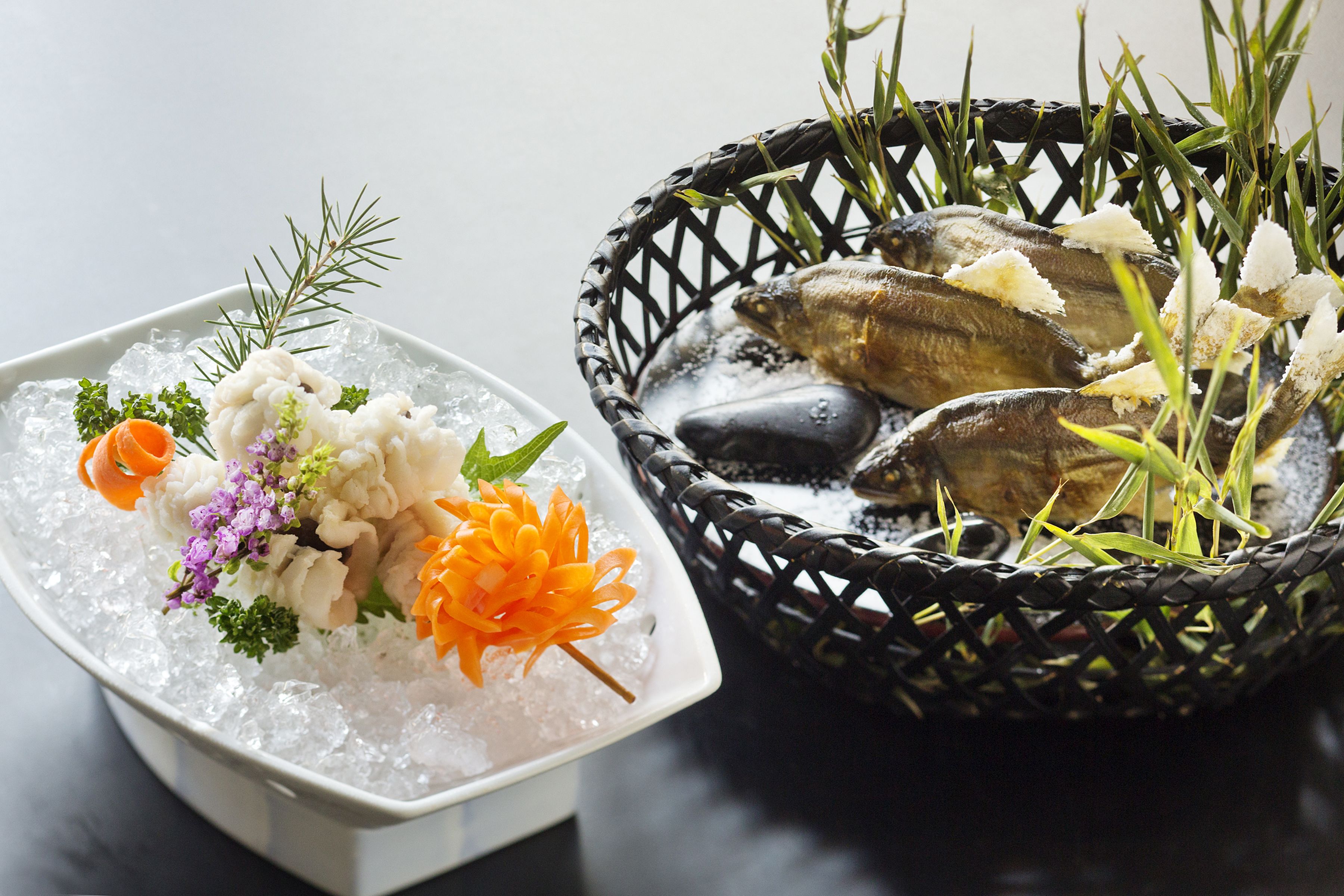 A dish of seafood and vegetables on ice, garnished with herbs and flowers, next to a woven basket with more seafood and greenery.