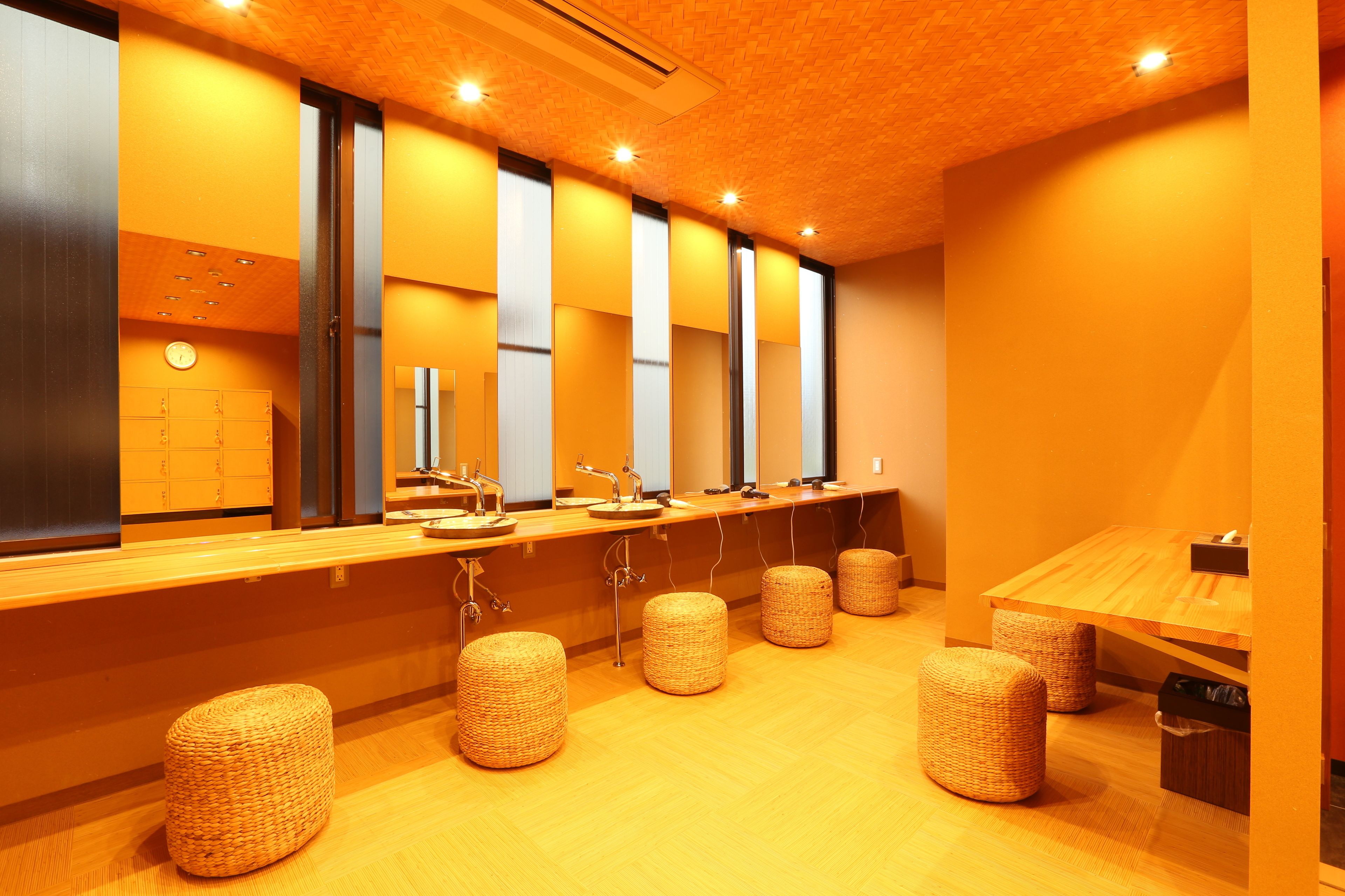 A row of sinks with mirrors, wicker stools, and a wooden table in a brightly lit bathroom.