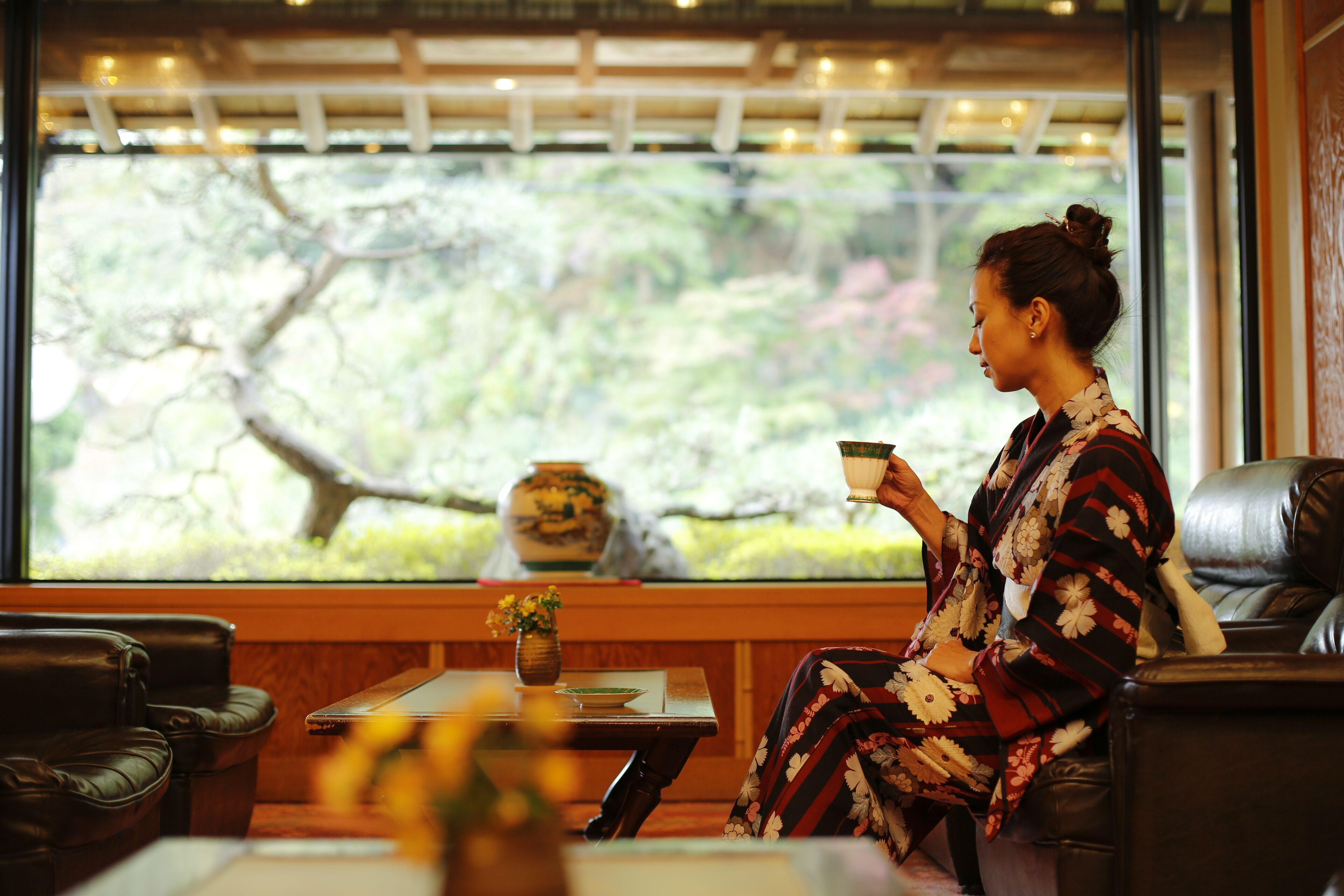A woman sitting by a window with a view of trees, a vase with flowers, and a table with a teapot.