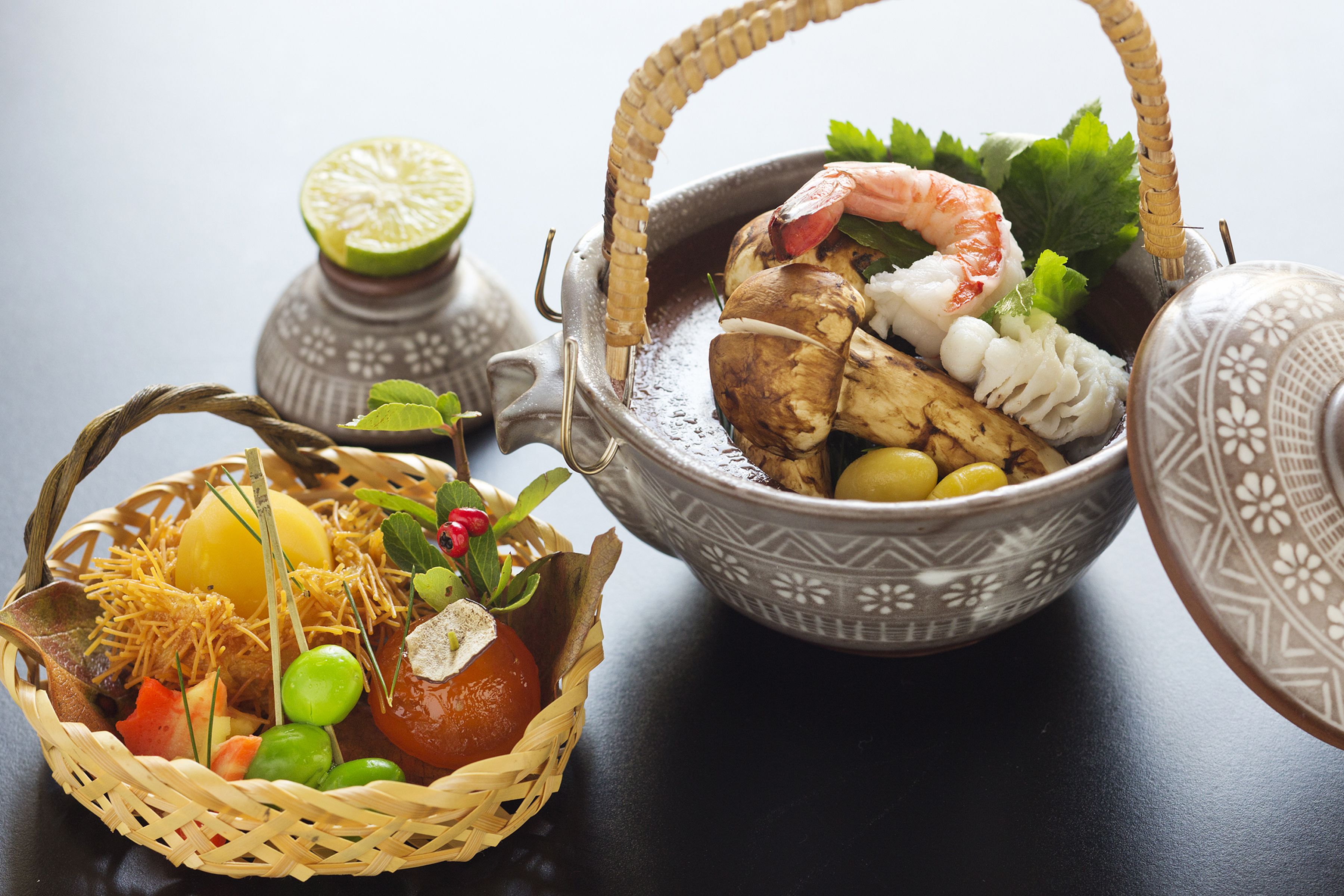 A table with a woven basket containing fruits and vegetables, and a ceramic bowl with seafood and herbs.