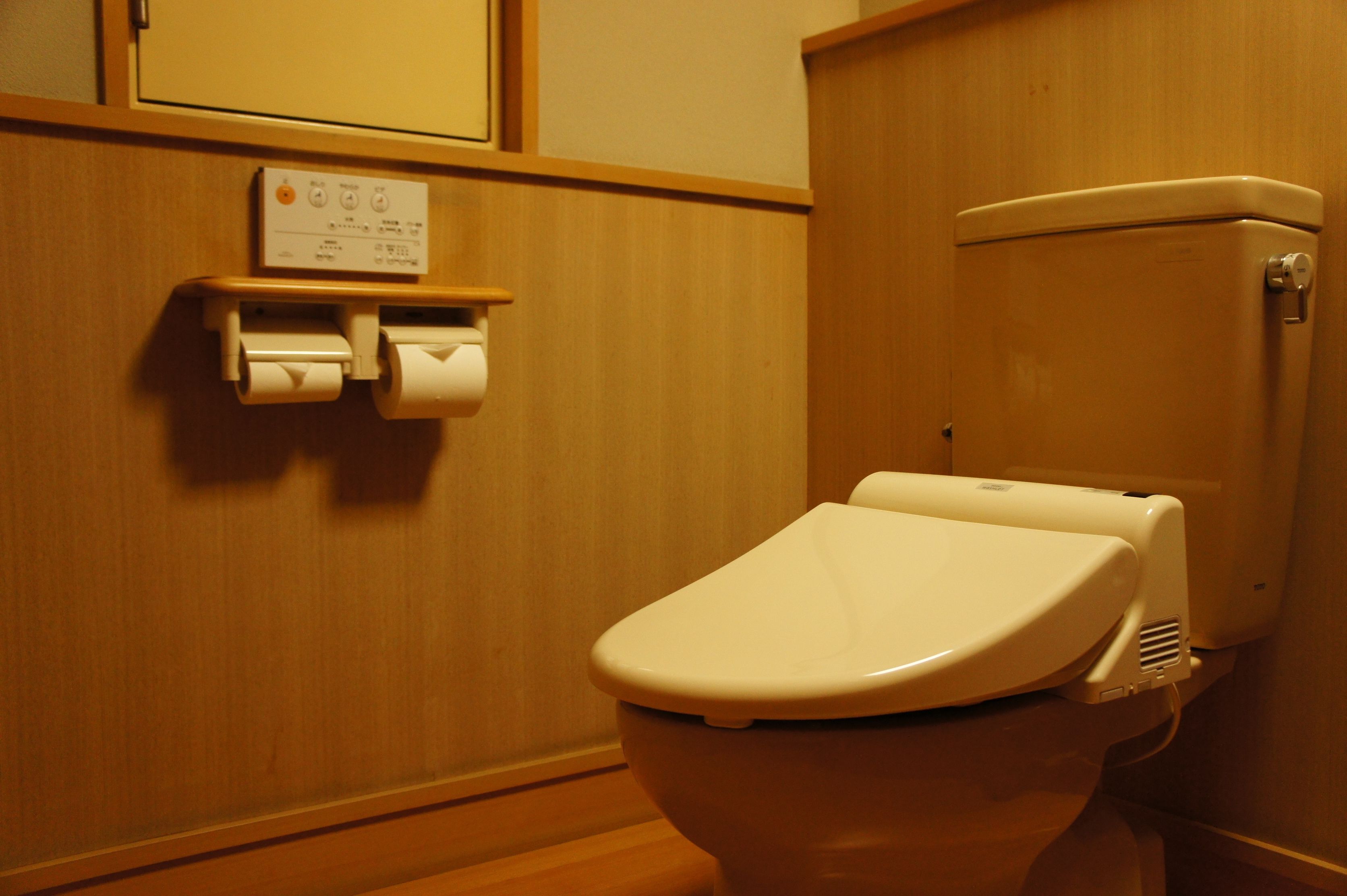 Inodoro con asiento y tapa blancos, dispensador de toallas de papel y palanca de descarga a la derecha.