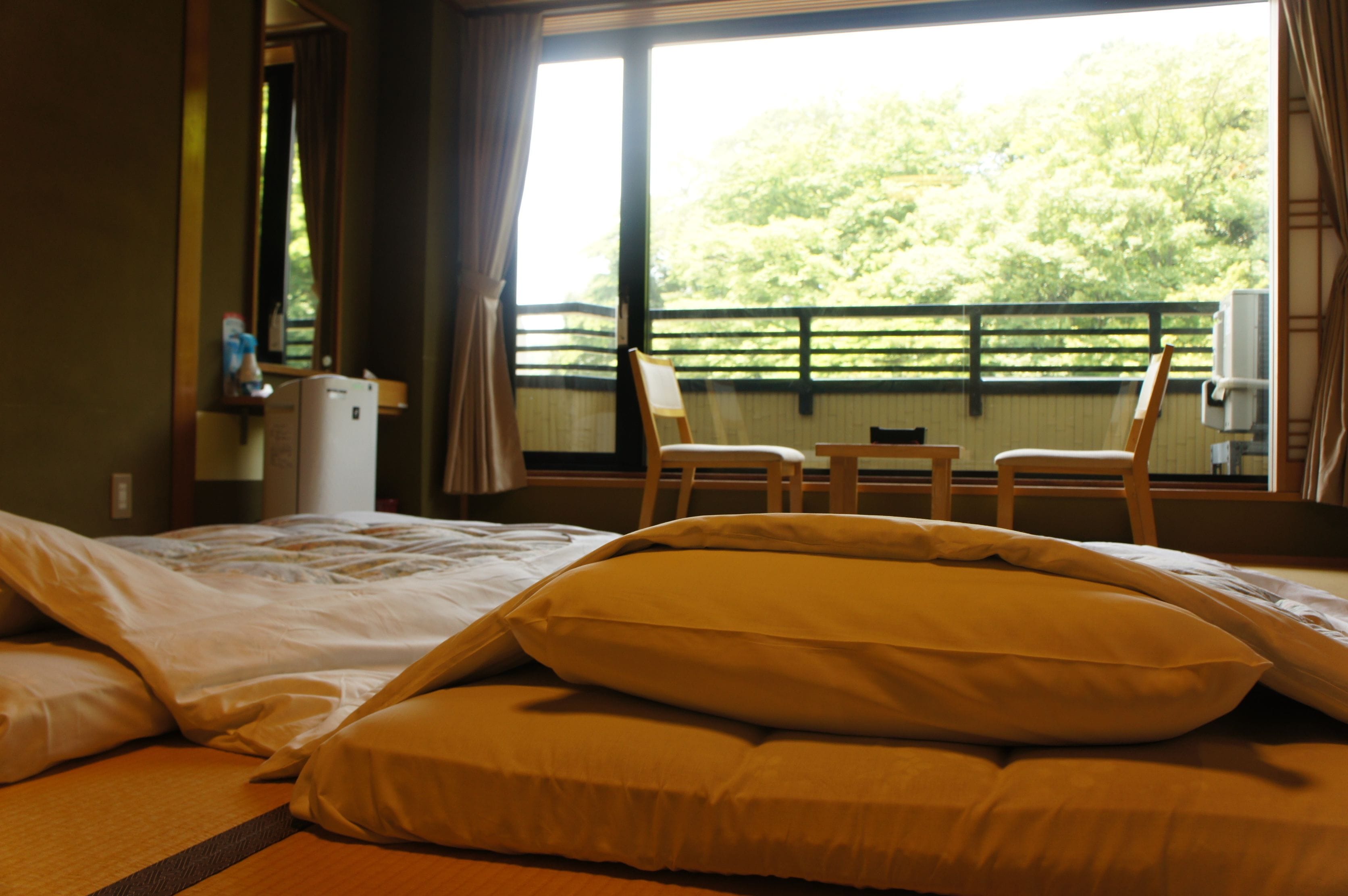 A hotel room with a bed, pillows, a small table, chairs, and a window with a view of trees.