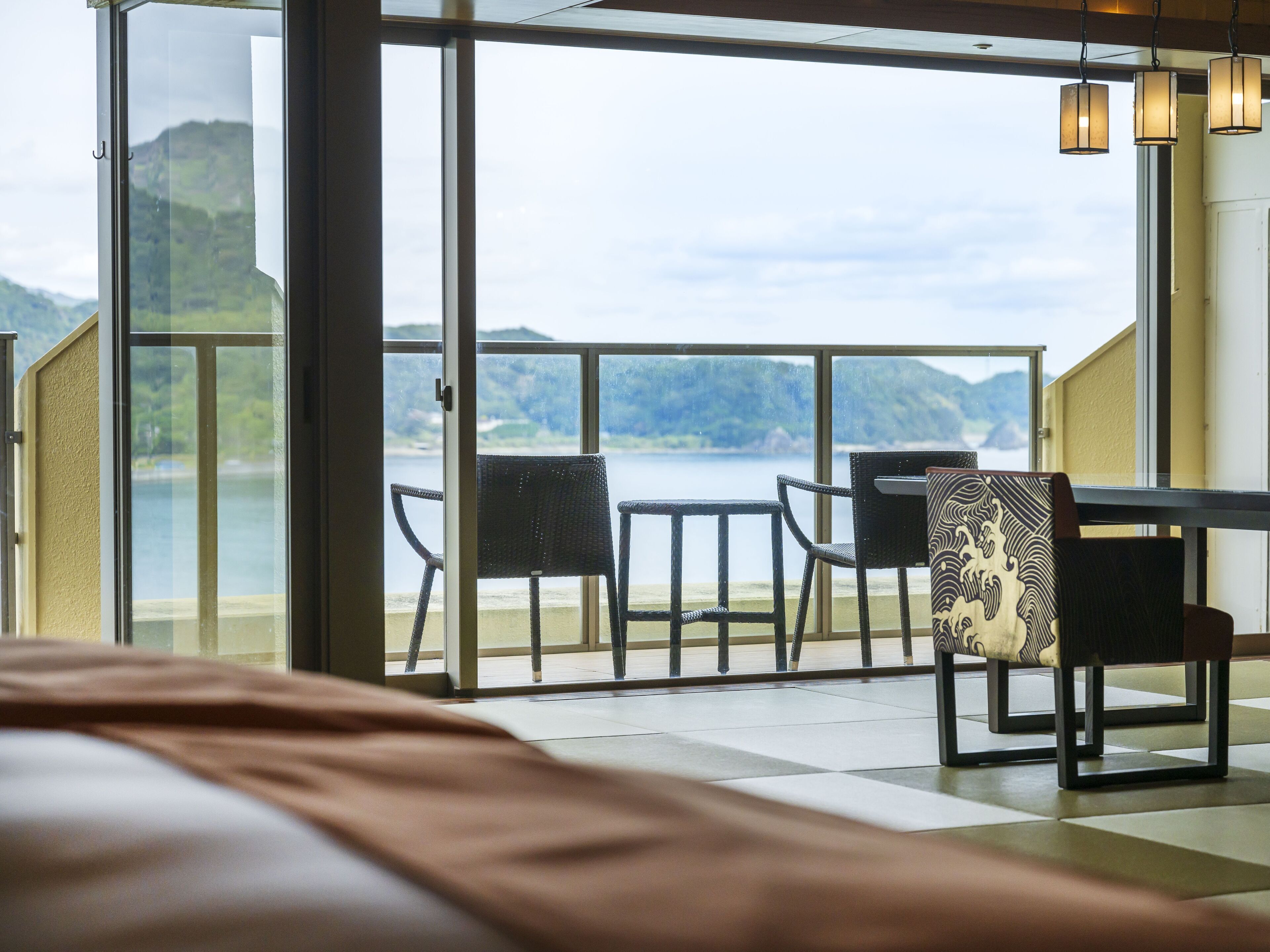 A balcony with a table and chairs overlooking a lake, with a bed in the foreground.