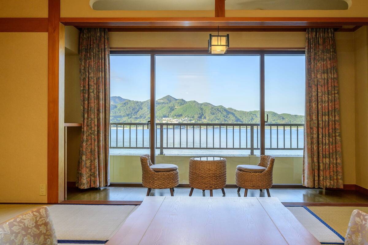 Ein Zimmer mit einem großen Holztisch, zwei Korbstühlen und Blick auf einen See und Berge durch große Fenster.