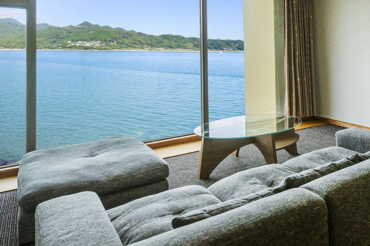 Ein Hotelzimmer mit großem Fenster und Blick auf einen See und Berge. Es gibt einen Glastisch und zwei grau gepolsterte Sessel.
