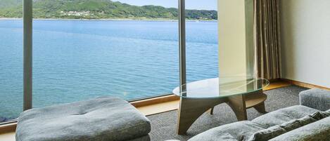 A hotel room with a large window offering a view of a lake and mountains. There is a glass table and two grey upholstered chairs.