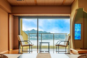 Ein Zimmer mit einer großen Schiebetür, die auf einen Balkon mit Blick auf Berge und einen See führt. Im Zimmer befinden sich zwei Korbstühle und ein kleiner Tisch.