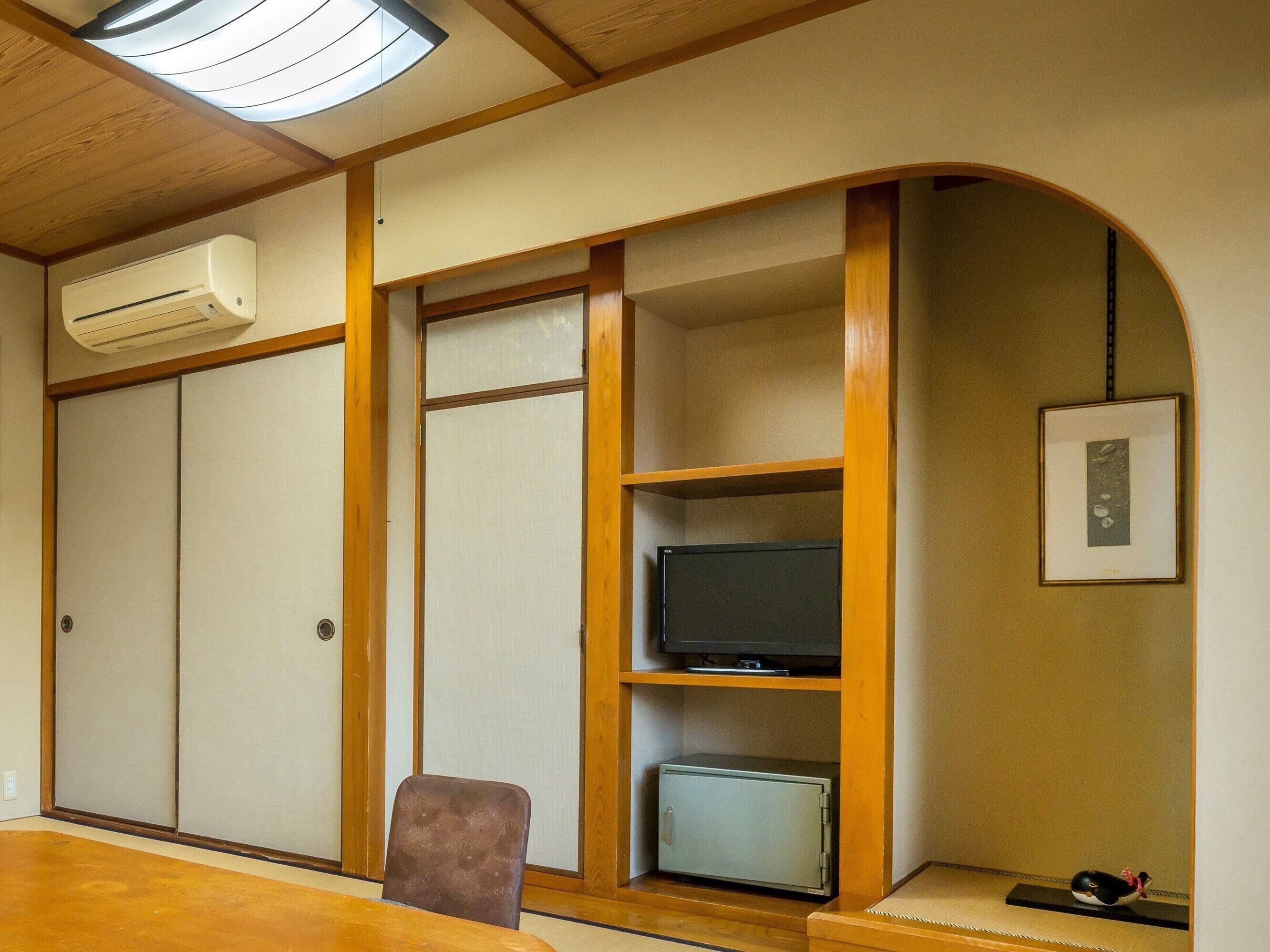 A room with a wooden ceiling, a built-in air conditioner, a television on a shelf, a small safe, and a framed picture on the wall.