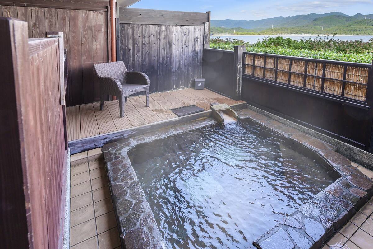 Ein Thermalbadbereich mit einer Holzterrasse, einem mit Steinplatten belegten Boden und Blick auf die Berge im Hintergrund.