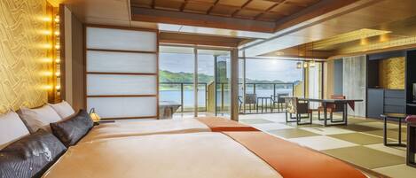 A modern hotel room with a large bed, a sofa, a dining area, and a view of the lake.