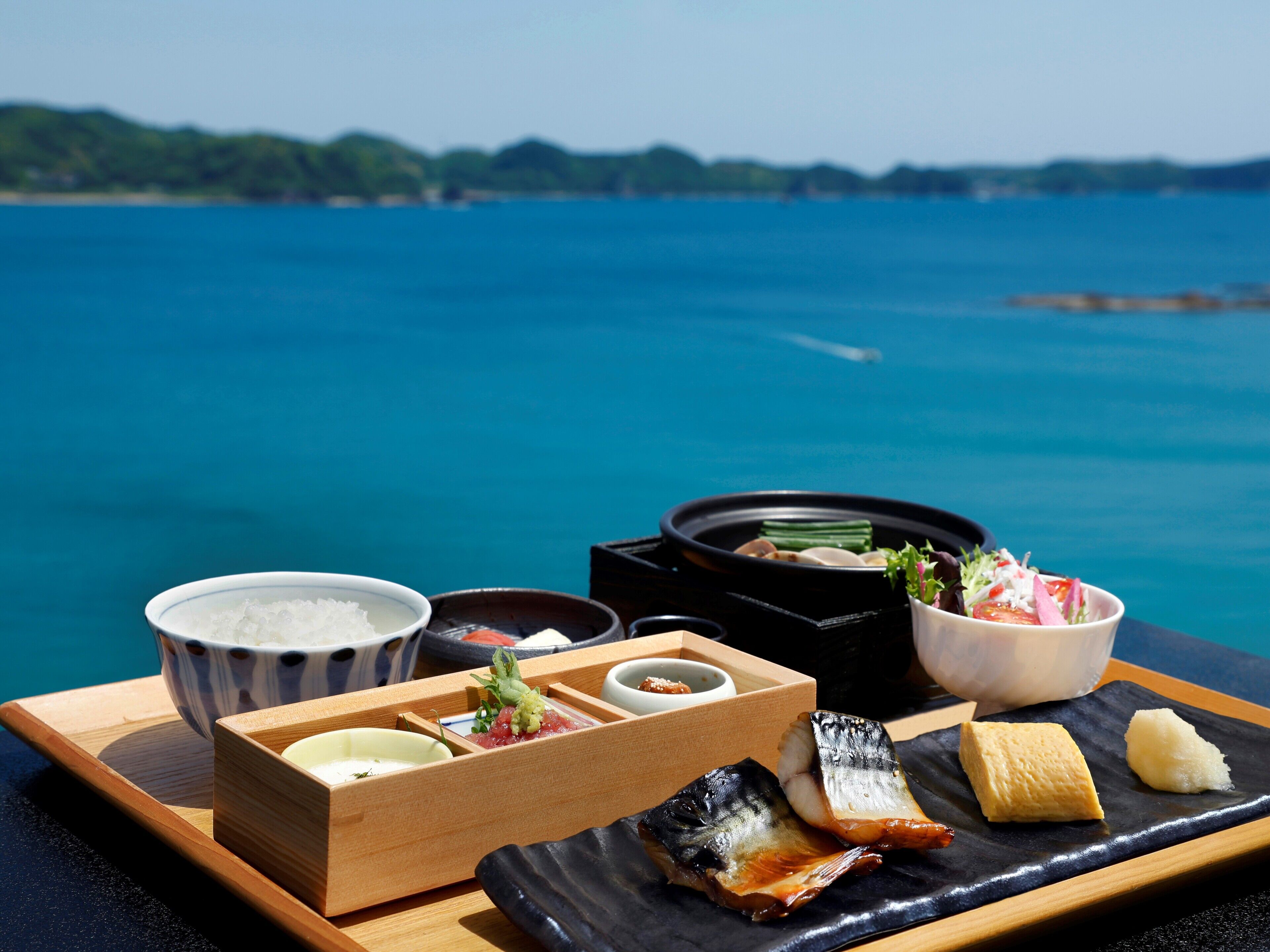 A wooden tray with various dishes including sashimi, a bowl of rice, and a small dish of sauce, set against a backdrop of a clear blue sea and distant hills.