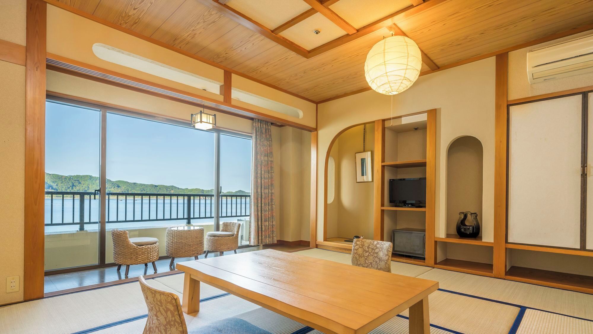 Ein traditioneller japanischer Raum mit Tatami-Bodenbelag, einem Holztisch, Korbstühlen, einem großen Fenster mit Seeblick und einem Deckenventilator.