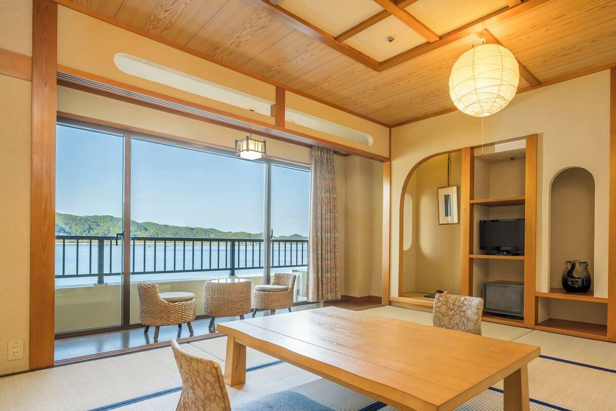 Ein traditioneller japanischer Raum mit Tatami-Bodenbelag, einem Holztisch, Korbstühlen, einem großen Fenster mit Seeblick und einem Deckenventilator.