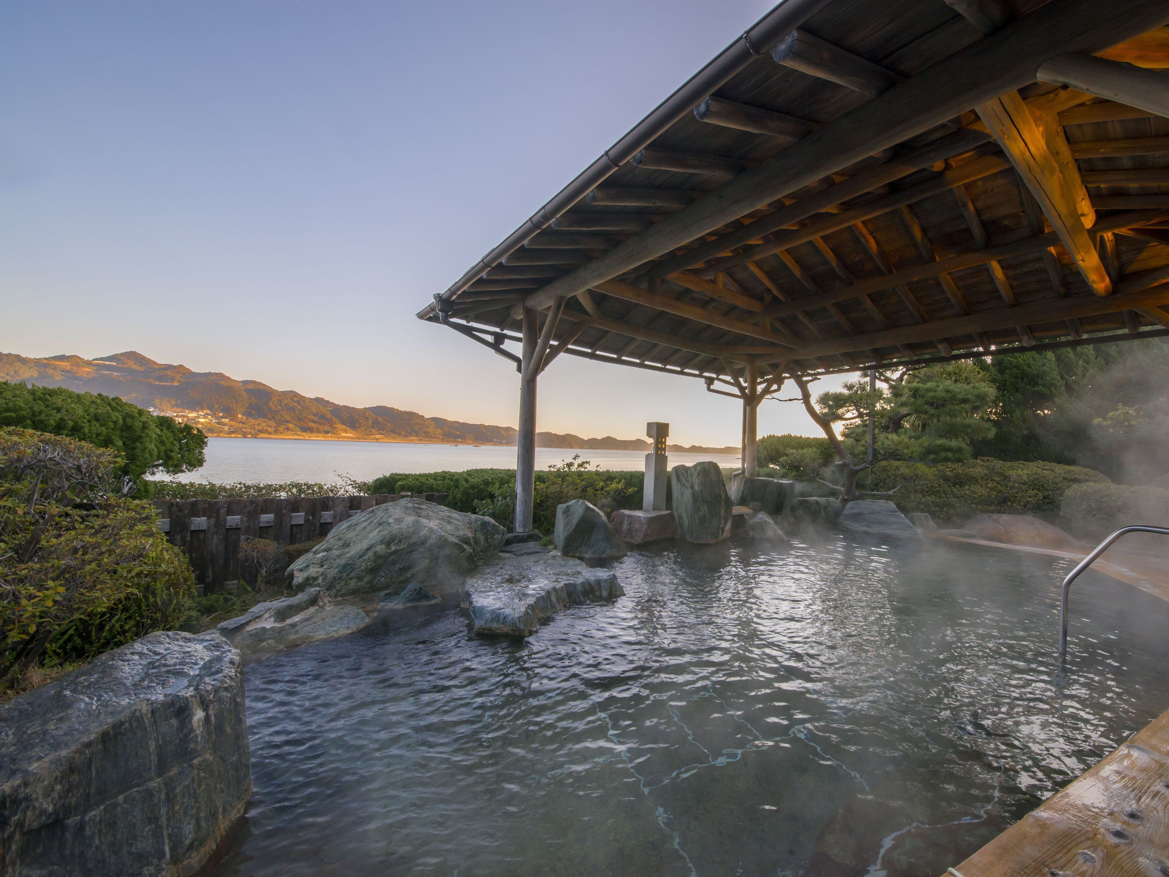 A traditional Japanese onsen with a wooden structure, rocky bath, and a view of mountains and water.