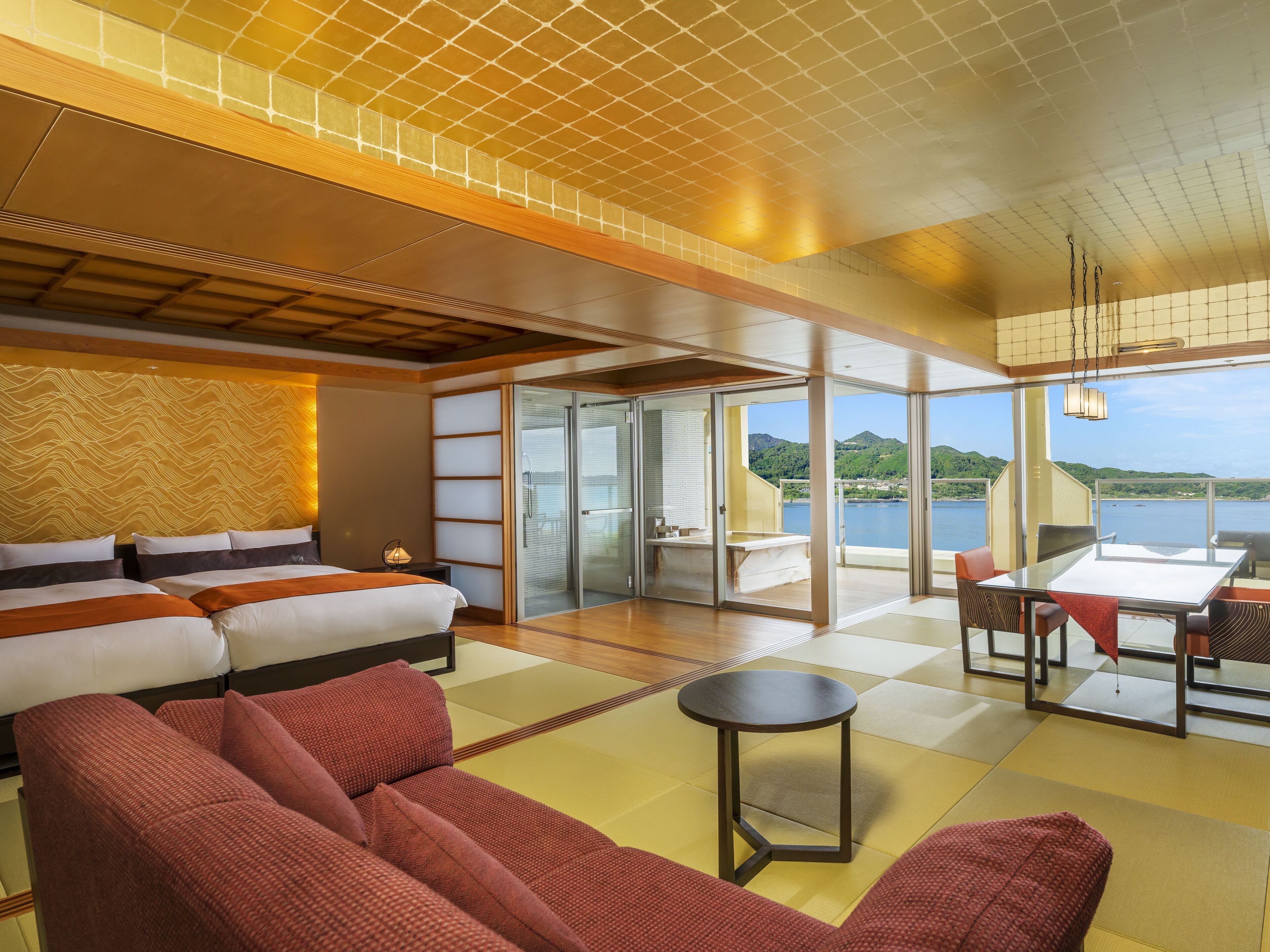 A modern hotel room with a large bed, a sitting area with red sofas, a dining table, and a view of a lake and mountains.