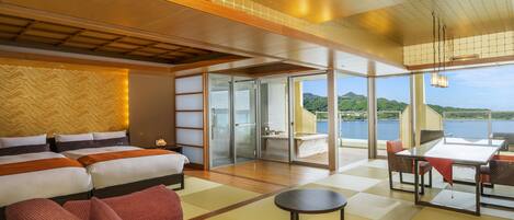 A modern hotel room with a large bed, a sitting area with red sofas, a dining table, and a view of a lake and mountains.