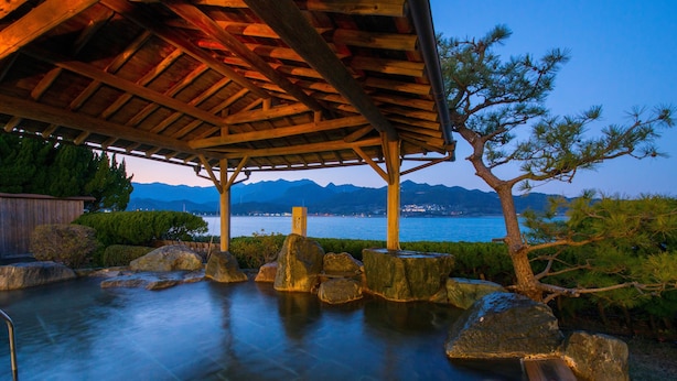 A traditional Japanese onsen with a wooden structure, large rocks, and a view of mountains and water.