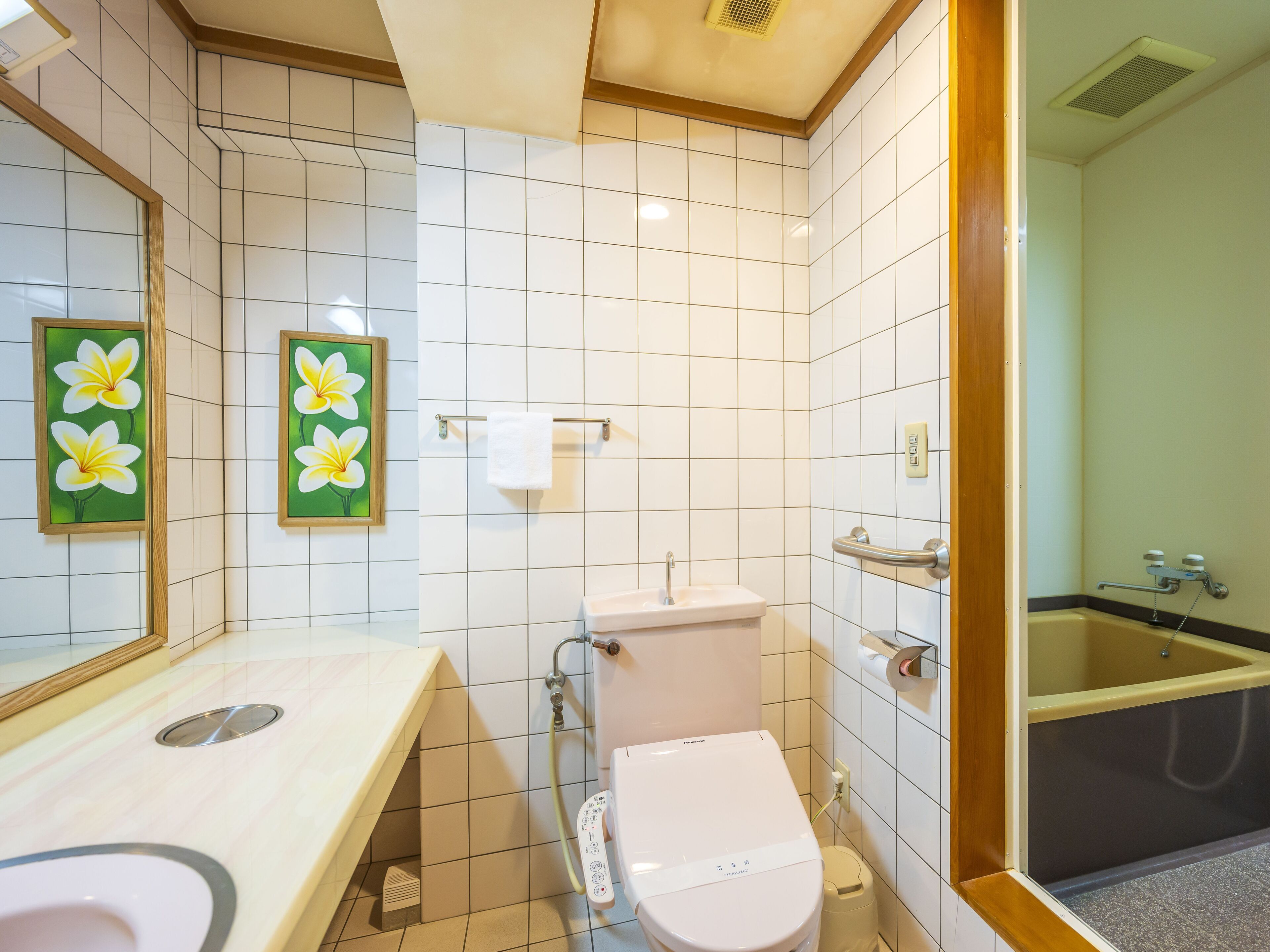 Een badkamer met witte tegels, een toilet, een wastafel, een douche, en twee bloemschilderijen in lijsten aan de muur.