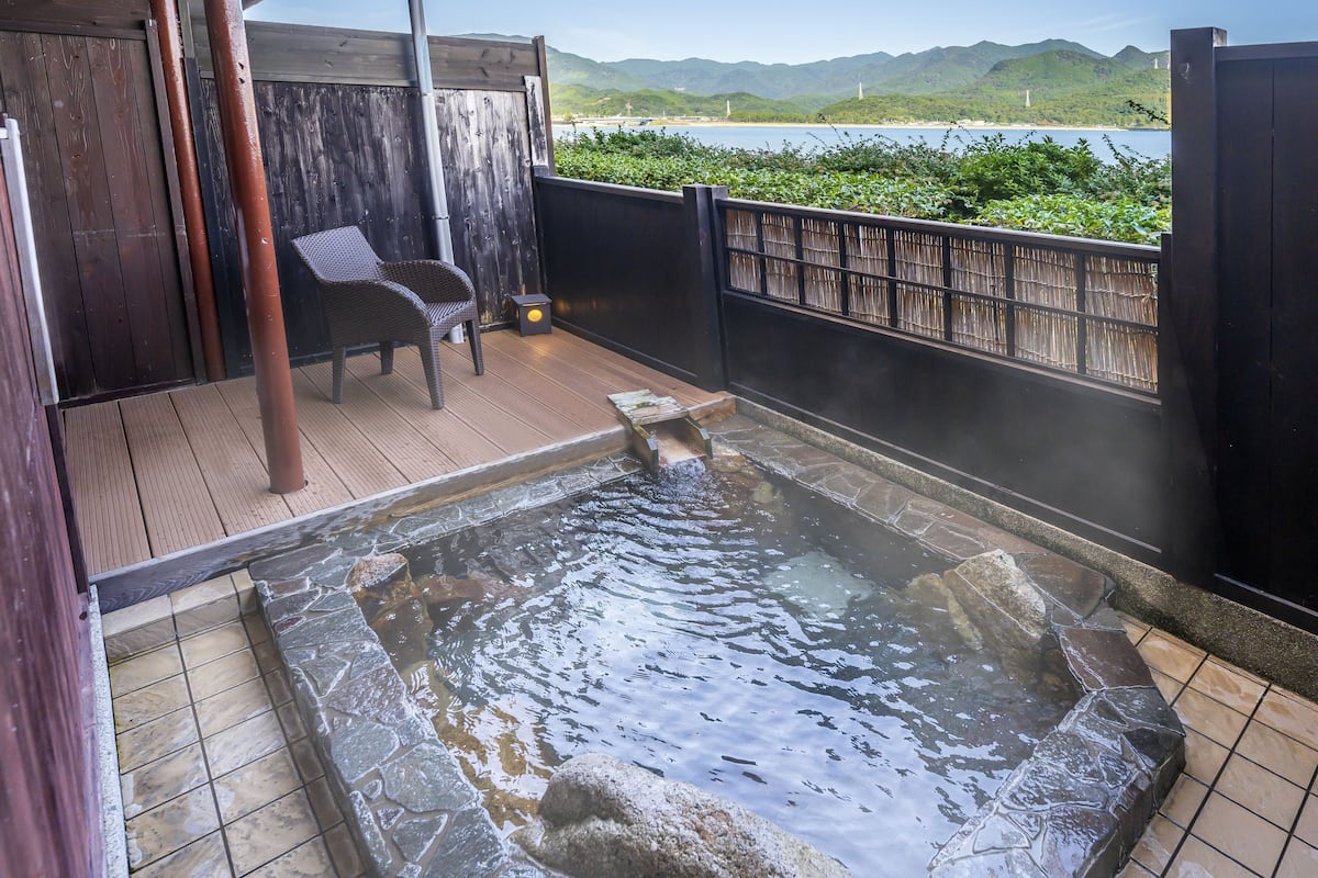 Ein Thermalbad mit Holzterrasse, Steinplattenboden und Blick auf Strand und Berge im Hintergrund.