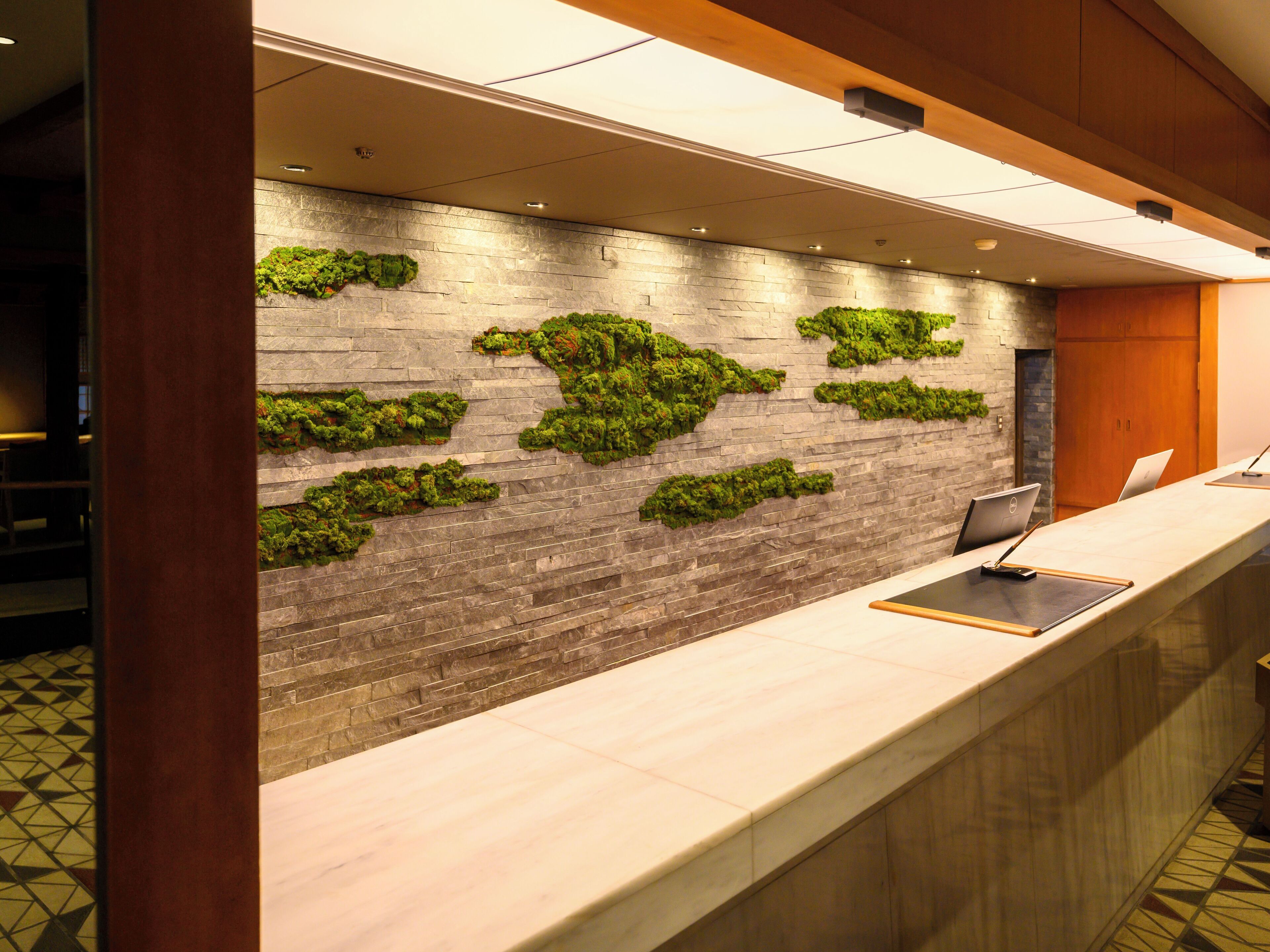 Un mostrador de recepción con pared de piedra y decoración de musgo verde, una computadora portátil y un teléfono inteligente sobre el mostrador.