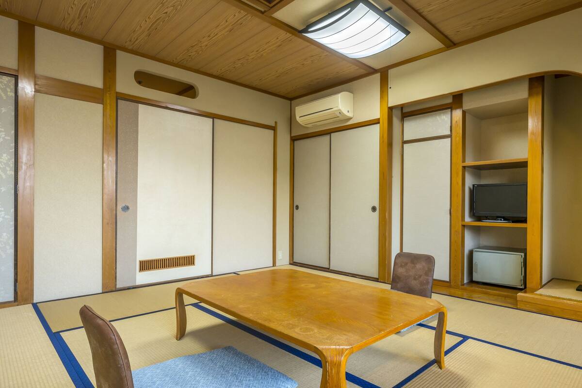 Ein traditioneller japanischer Raum mit Tatami-Böden, einem Holztisch, einem Fernseher und einem kleinen Kühlschrank.