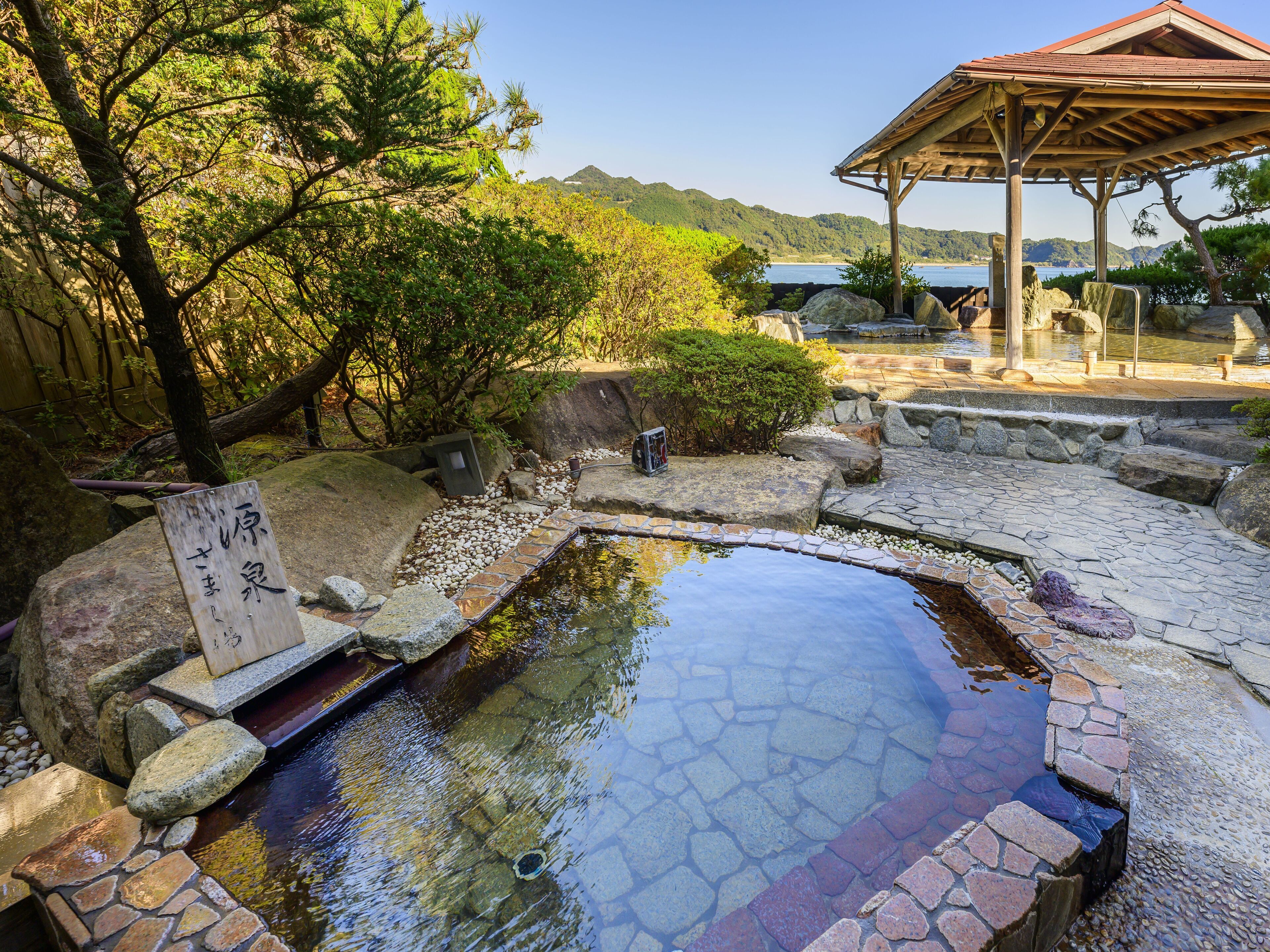 Un onsen japonais traditionnel, avec des marches en pierre menant à une source d’eau chaude, entouré d’une végétation luxuriante et agrémenté d’un pavillon en bois.