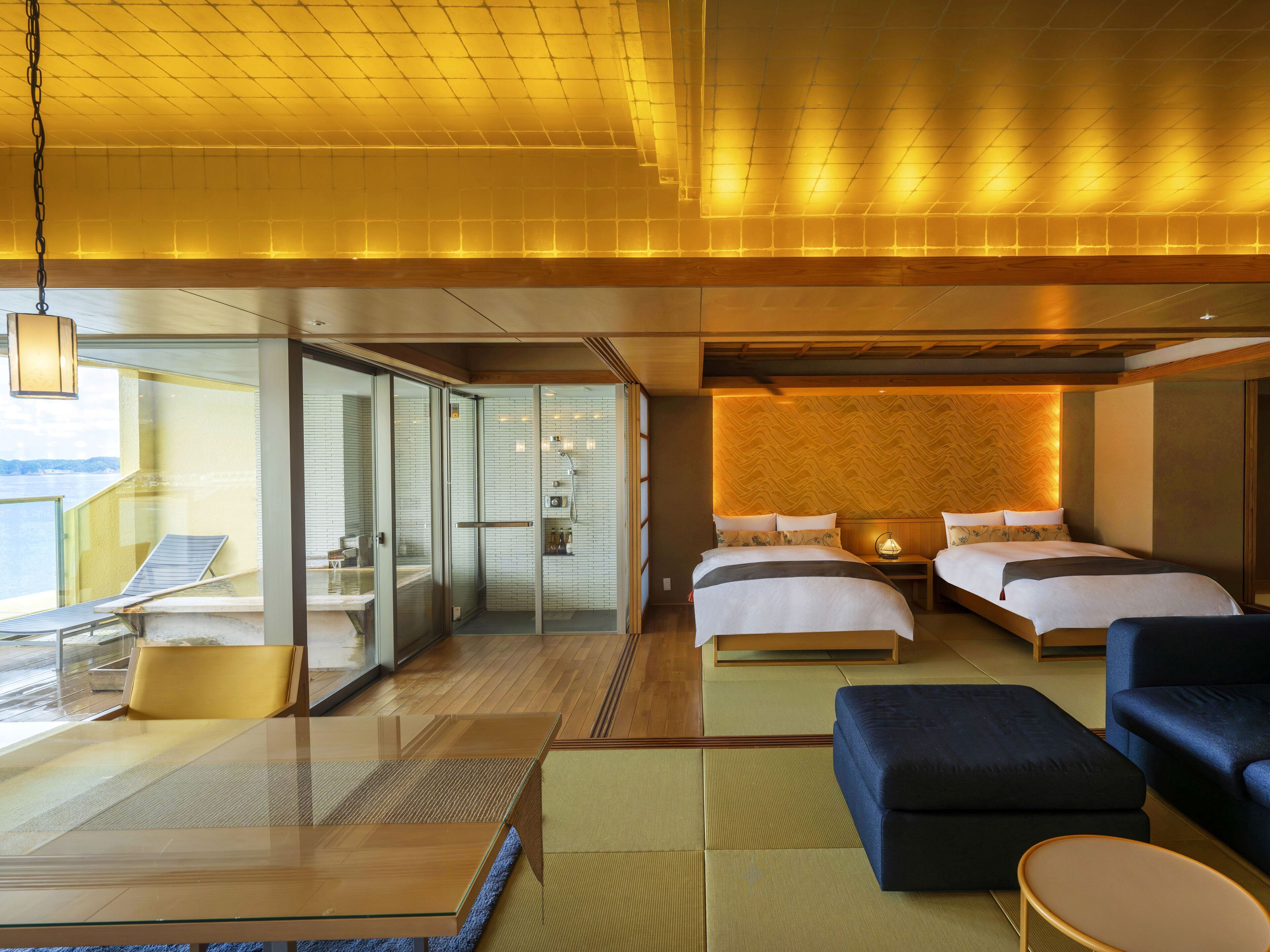 A modern hotel room with a glass-enclosed shower, two beds, a sitting area with a table, and a view of the sea.