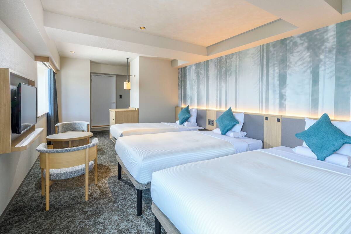 Ein Hotelzimmer mit zwei Betten, einem Schreibtisch, einem Stuhl, einem Fernseher und einem großen Fenster mit Blick auf einen Wald.