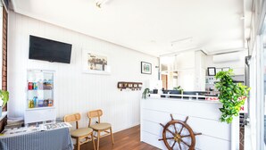 Una habitación con piso de madera, una mesa con periódicos, dos sillas de mimbre, un televisor montado en la pared, un refrigerador con bebidas, un adorno de timón de barco y una planta.