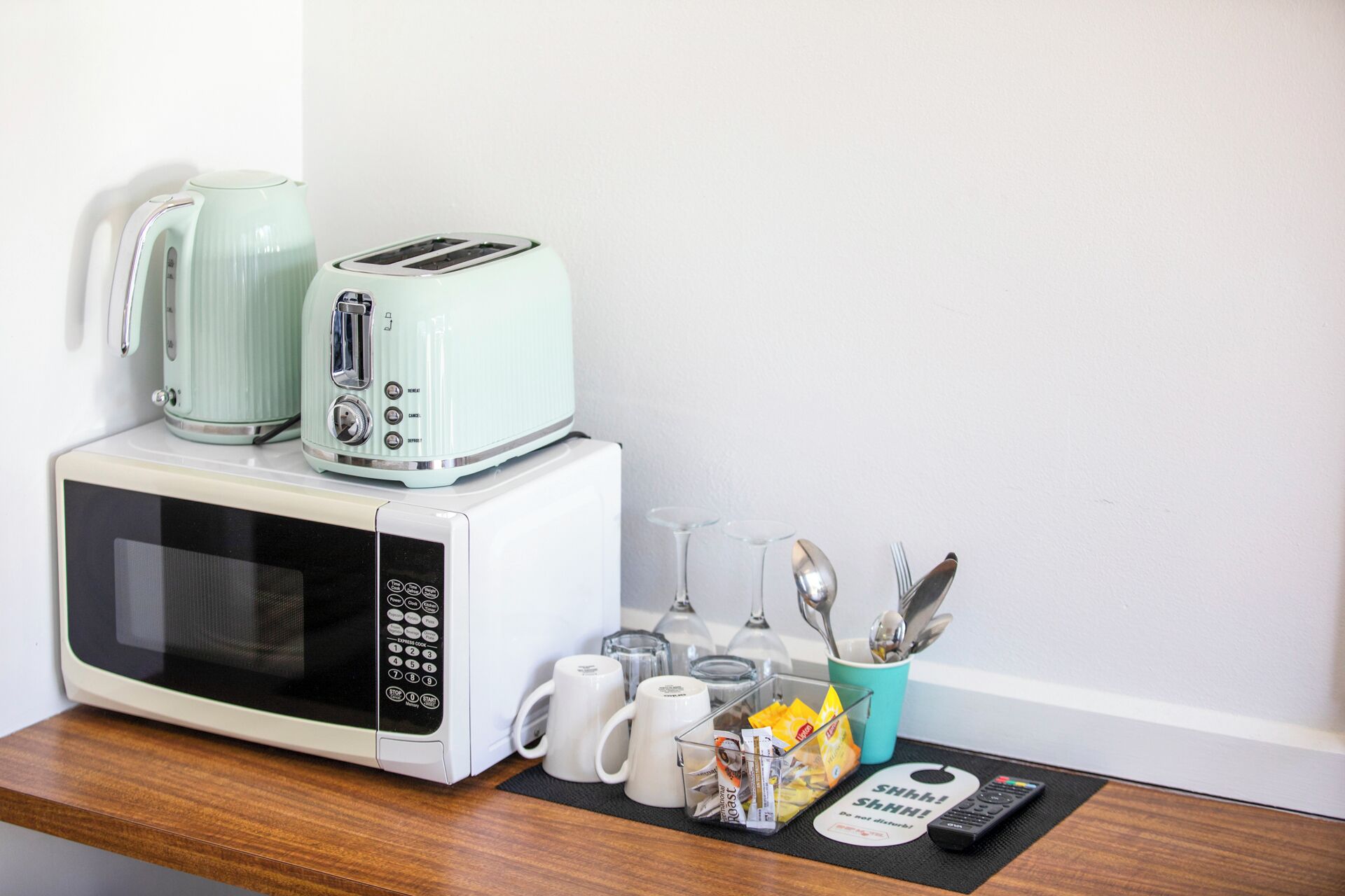 A kitchenette with a microwave, toaster, and kettle. There are utensils, a mug, and a remote control on the counter.