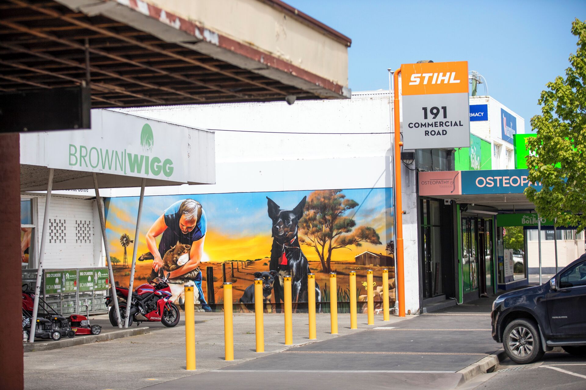 Zona comercial con un mural grande, una farmacia y un letrero que dice 'STIHL 191 COMMERCIAL ROAD'.