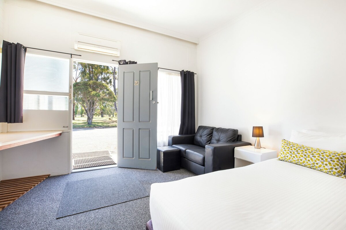 Hotelkamer met een bed, een bureau, een stoel, een nachtkastje en een deur naar buiten.