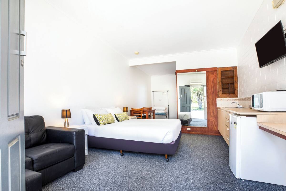 A compact hotel room with a bed, a small desk, a chair, a TV, and a kitchenette.