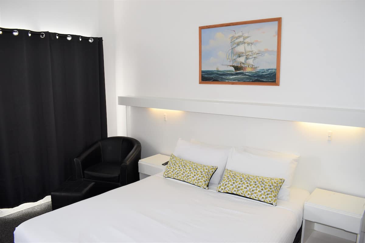 A hotel room with a bed, a chair, a nightstand, a painting of a ship on the wall, and a black curtain.