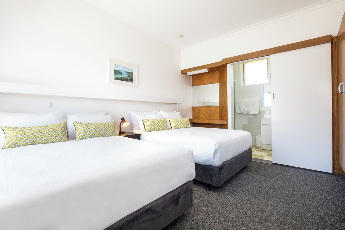 Een hotelkamer met twee bedden, een klein nachtkastje met een lamp, een houten hoofdbord, een spiegel en een badkamer die door een open deur zichtbaar is.