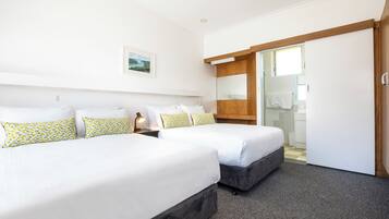 A hotel room with two beds, a small bedside table with a lamp, a wooden headboard, a mirror, and a bathroom visible through an open door.
