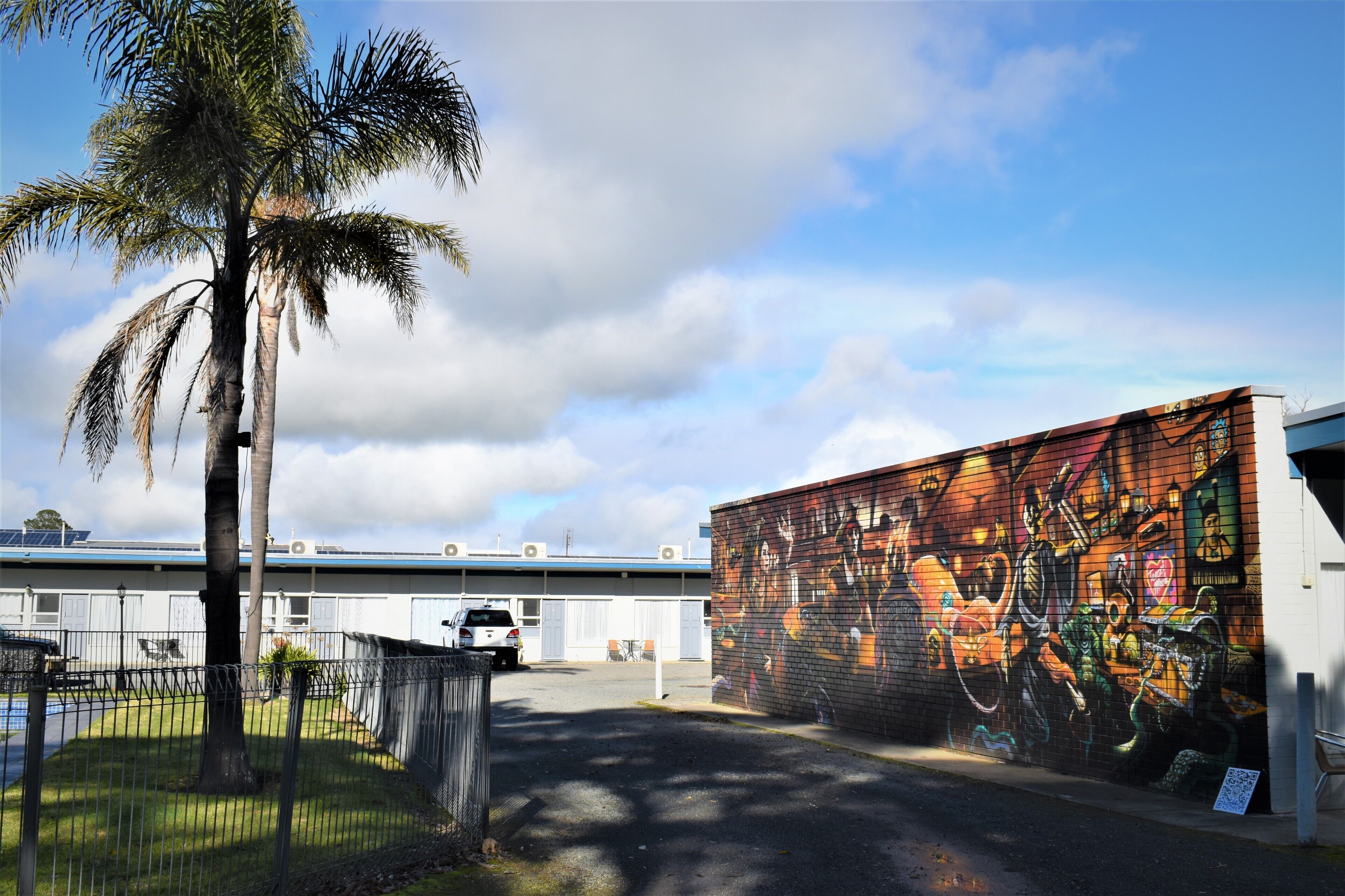 Un estacionamiento con un mural en un edificio, una palmera y una zona delimitada por una cerca.