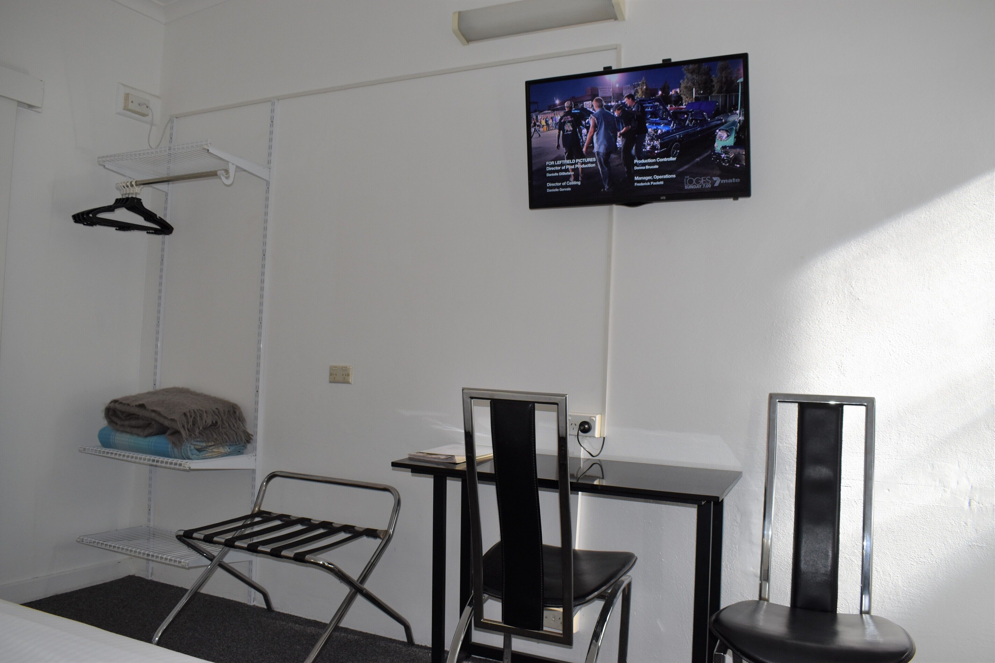 Habitación de hotel con televisor montado en la pared, un escritorio con silla, una mesita pequeña y un perchero.