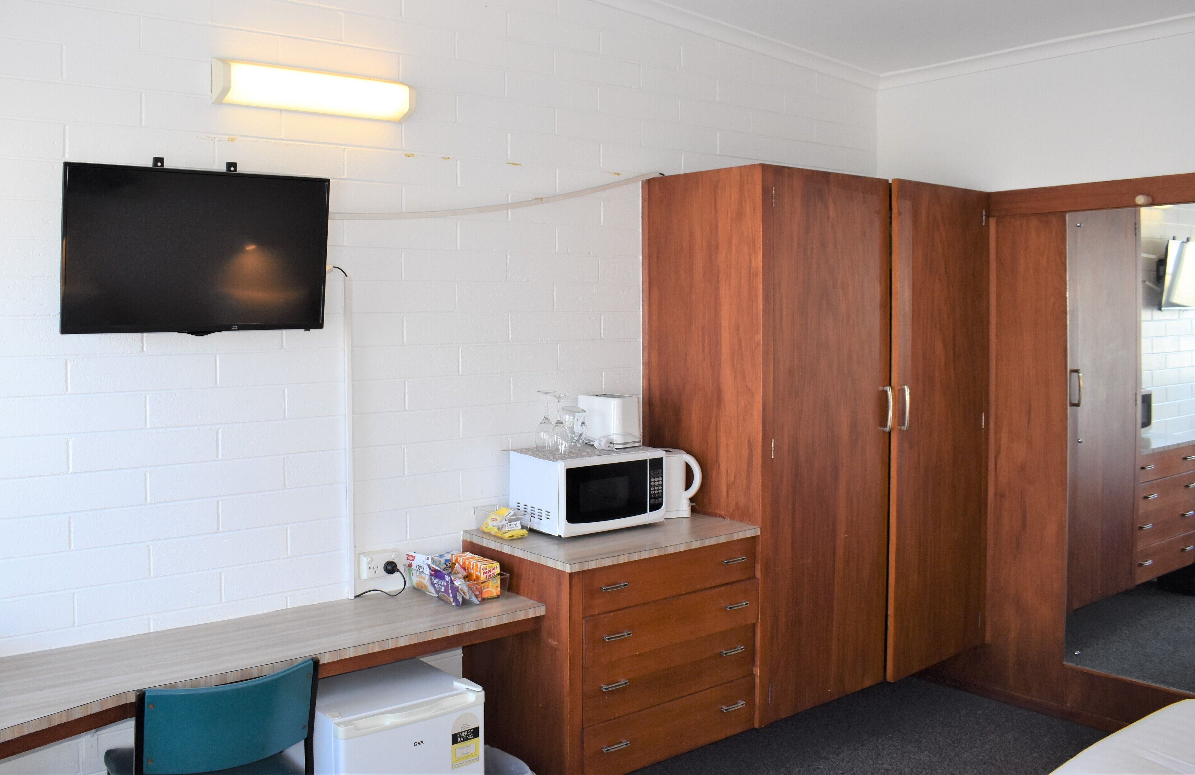 Une pièce compacte avec un téléviseur à écran plat fixé à un mur en briques blanches, une armoire en bois avec plusieurs tiroirs, un petit coin cuisine équipé d’un four à micro-ondes et une seule chaise.