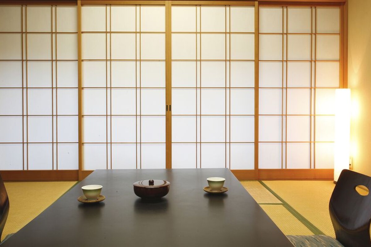 Ein traditioneller japanischer Raum mit Tatami-Böden, einem niedrigen Tisch mit einer Schüssel und zwei Tassen sowie Schiebetüren im Hintergrund.