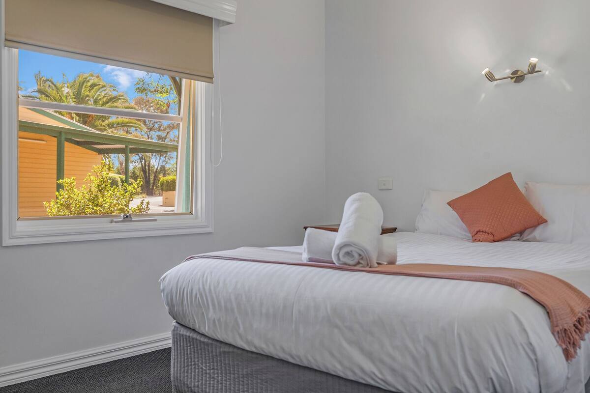 A hotel room with a bed, a window with a view of palm trees, a wall-mounted light, and a small table with a towel.