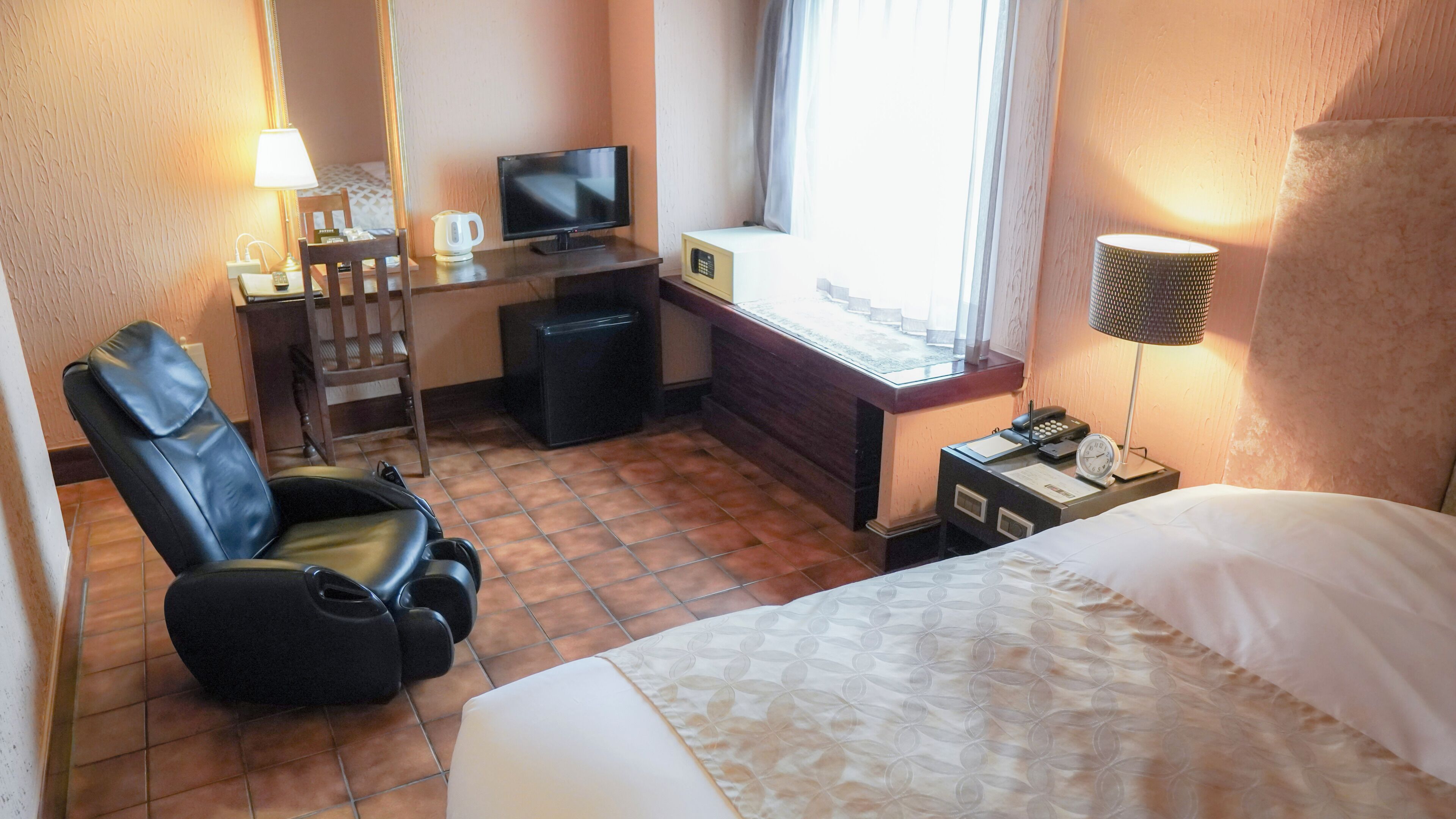 A hotel room with a bed, a desk with a television, a chair, a small table with a lamp, and a window with curtains.