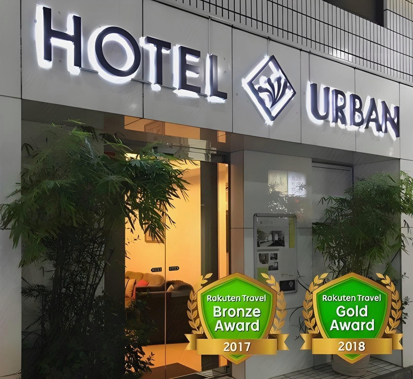 A hotel named 'HOTEL URBAN' with a modern design, including a glass entrance and indoor plants. It displays awards from 'Rakuten Travel' for Bronze in 2017 and Gold in 2018.