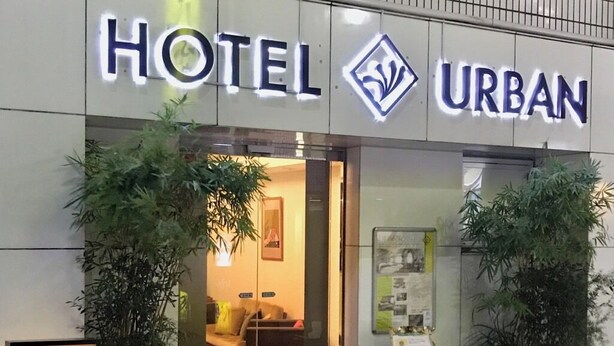 A hotel entrance with a sign reading 'HOTEL URBAN,' a reception desk, and potted plants.