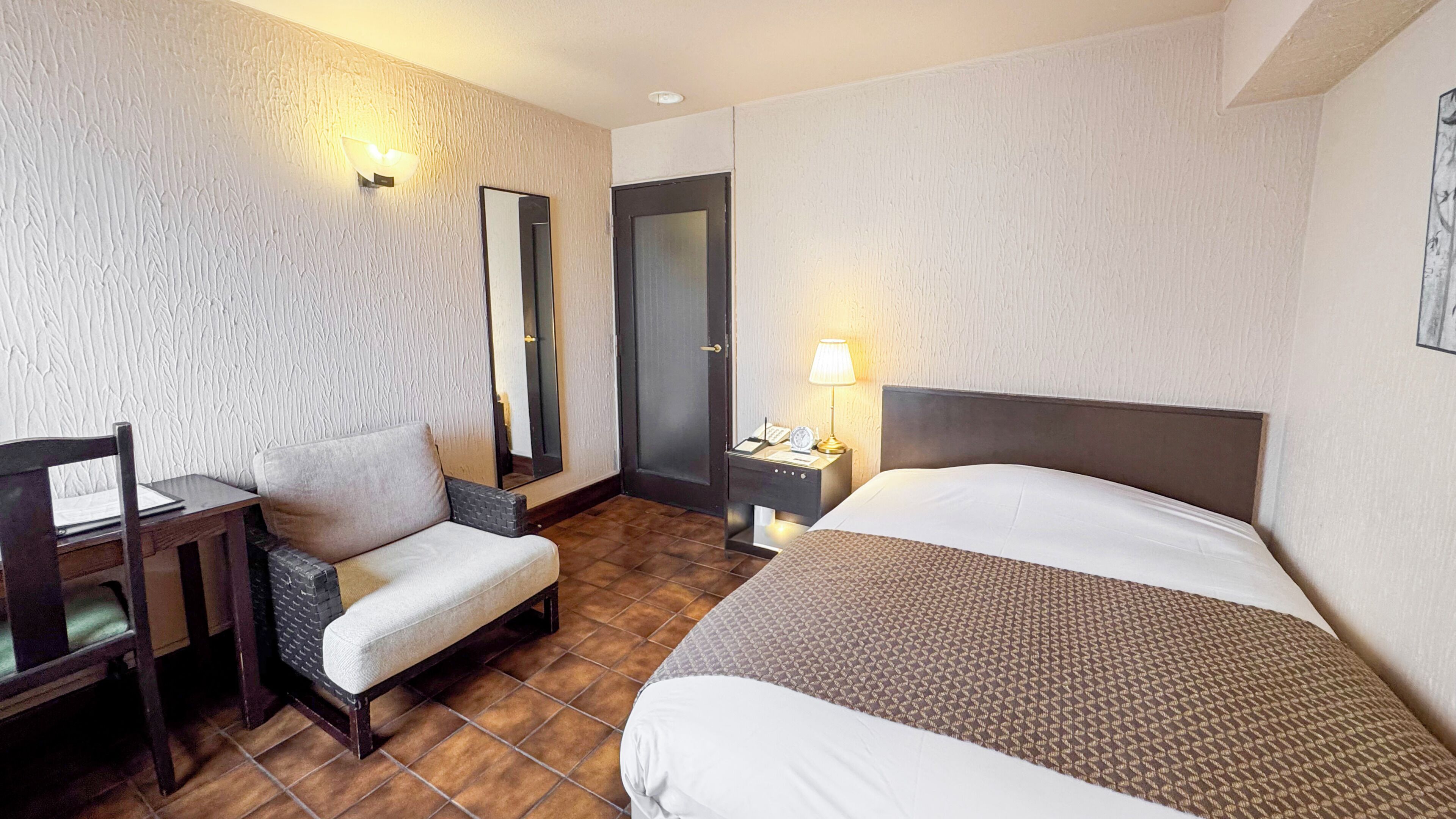 A hotel room with a bed, a chair, a desk, a nightstand, a lamp, and a mirror.