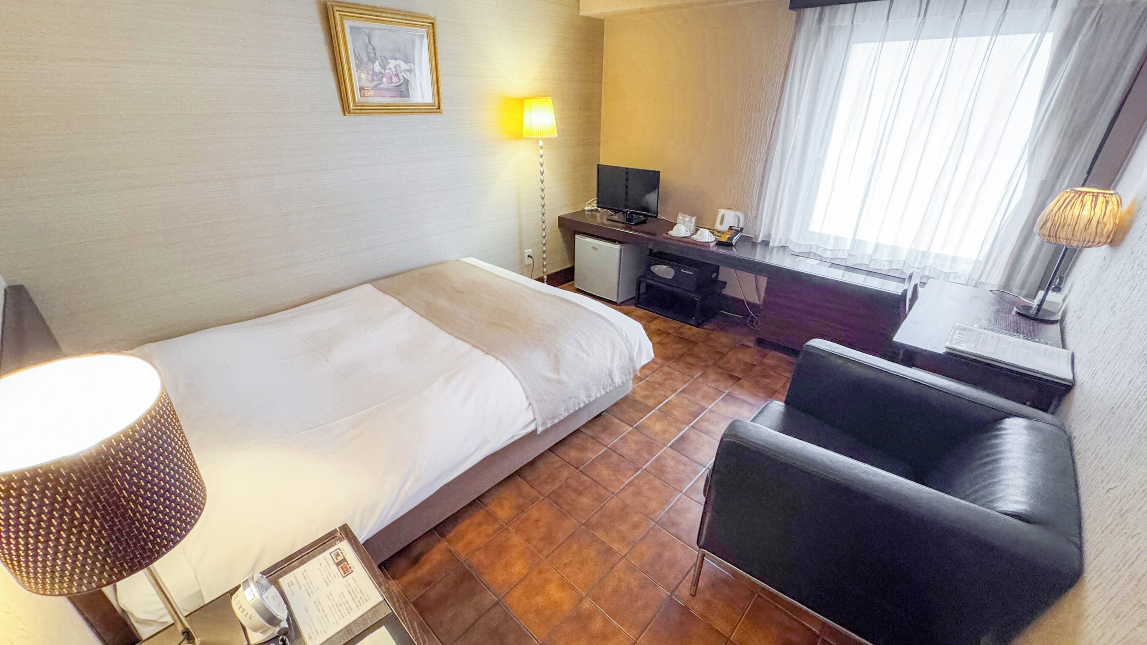 A hotel room with a bed, a desk, a chair, a lamp, and a window with curtains.
