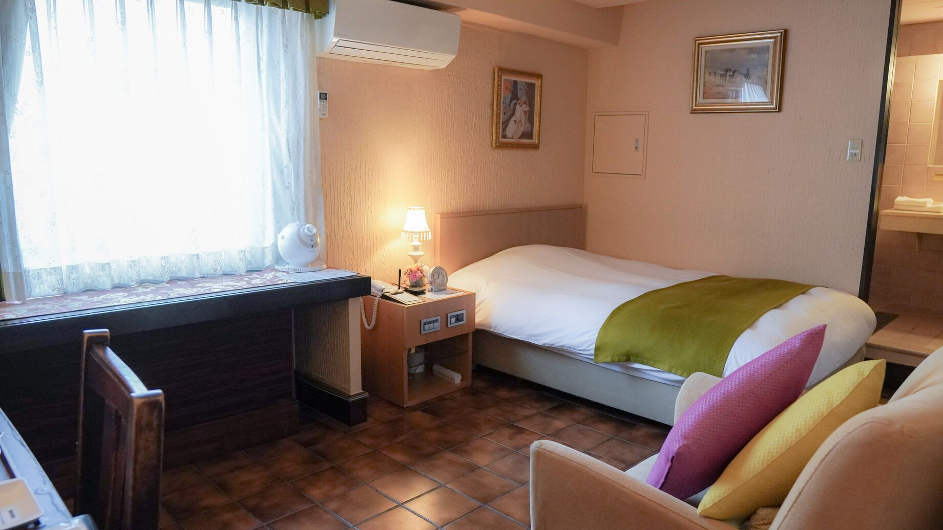 Ein kleines Hotelzimmer mit einem Bett, einem Schreibtisch mit Lampe, einem Stuhl, einem Fenster mit Vorhängen und einem Bild an der Wand.